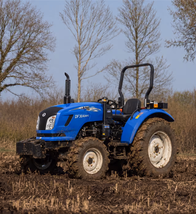 Plavi traktor DF304MC sa kabinom i velikim blatnjavim točkovima stoji na oranoj zemlji, sa još jednom plitkom brazdom u prvom planu. Gole grane drveća i bledo nebo u pozadini stvaraju ruralnu, zimsku scenu, dok prirodni dnevni svetla naglašavaju zemljane tonove i metalnu površinu.