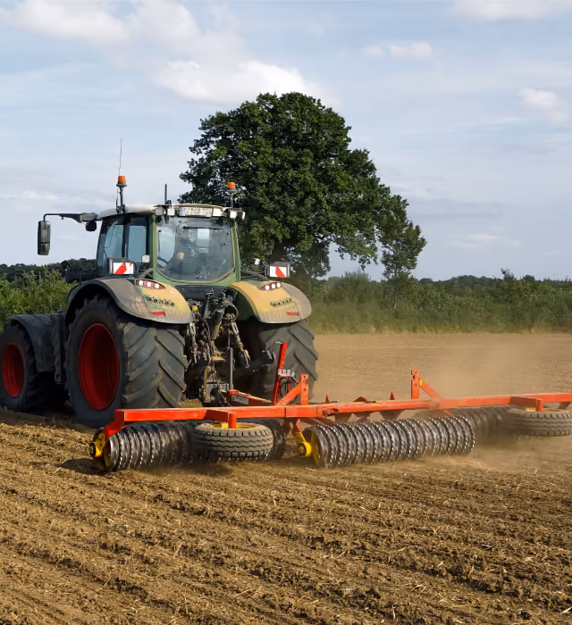 Veliki traktor izorava i ravna suvo polje pomoću širokog crvenog priključka sa valjcima, dok se za njim pružaju uredne brazde. U pozadini stoje usamljeno drvo i nizak oblak prašine pod vedrim, svetlim nebom.