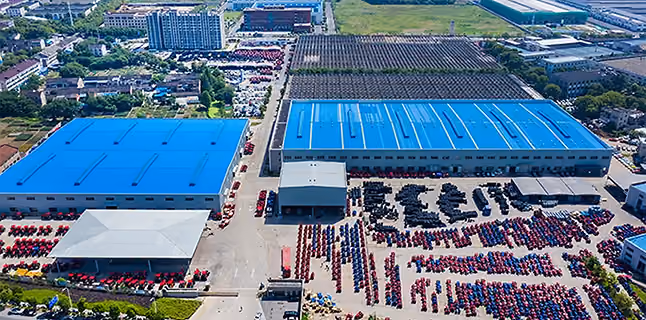 Pogled iz vazduha na veliki industrijski kompleks sa dve plave hale, belim šatorom i uredno poređanim redovima crvene i plave robe ili opreme u dvorištu. U pozadini se vide putevi, parkirani kamioni i zelene površine, pod jasnim dnevnim svetlom.