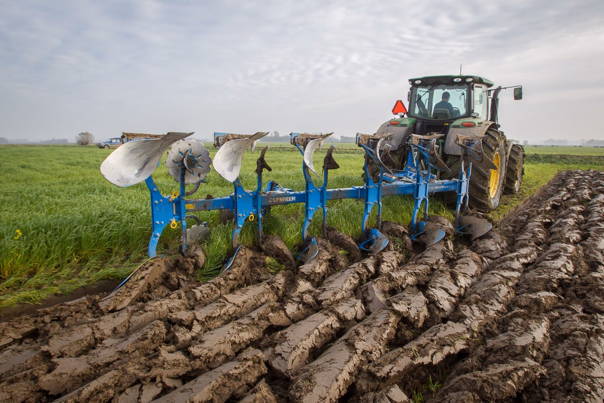 A tractor attached to a plow is tilling a muddy field next to green grass under a cloudy sky.