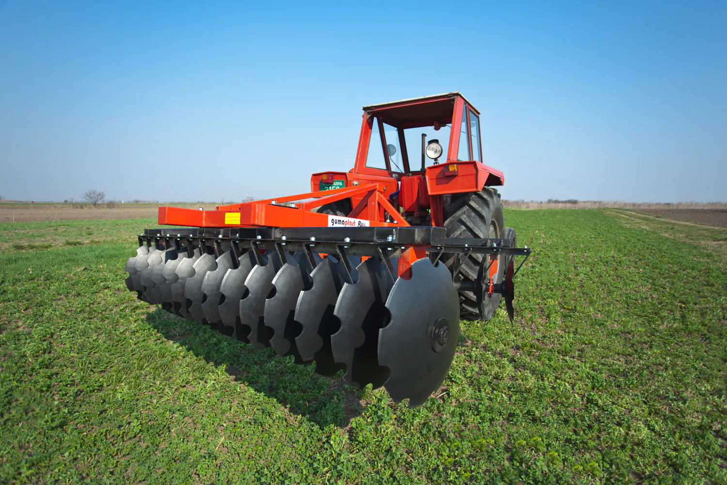 Red tractor equipped with a large black disc harrow attachment on a green farm field under clear blue sky.