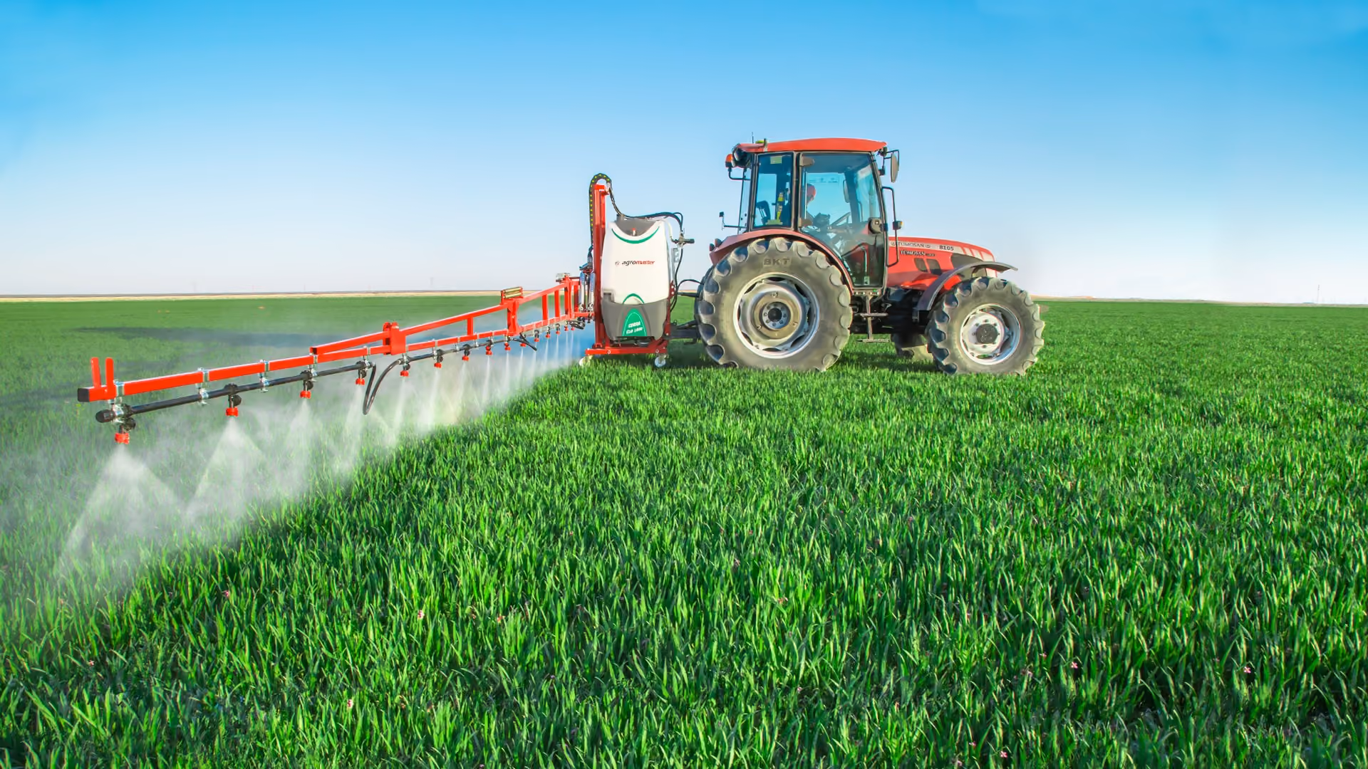 Red tractor equipped with an attached agricultural sprayer spraying mist over a lush green field under a clear blue sky.