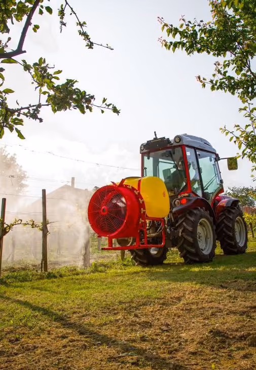 A red tractor with a yellow and red sprayer attachment spraying mist over an orchard or vineyard under a partly cloudy sky.