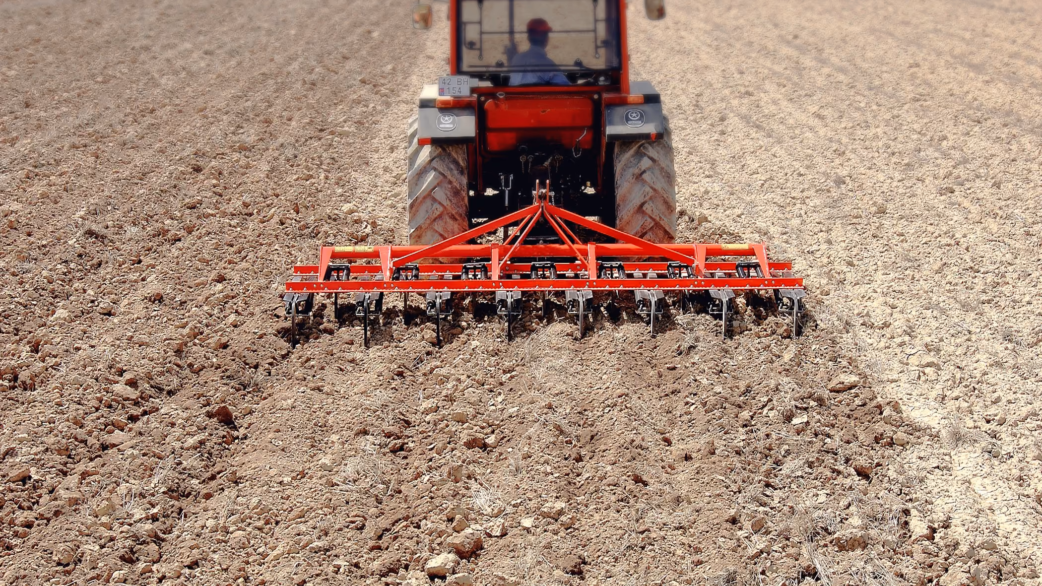 Red tractor with a cultivator attachment tilling and breaking up dry soil in a large open field from a rear view.