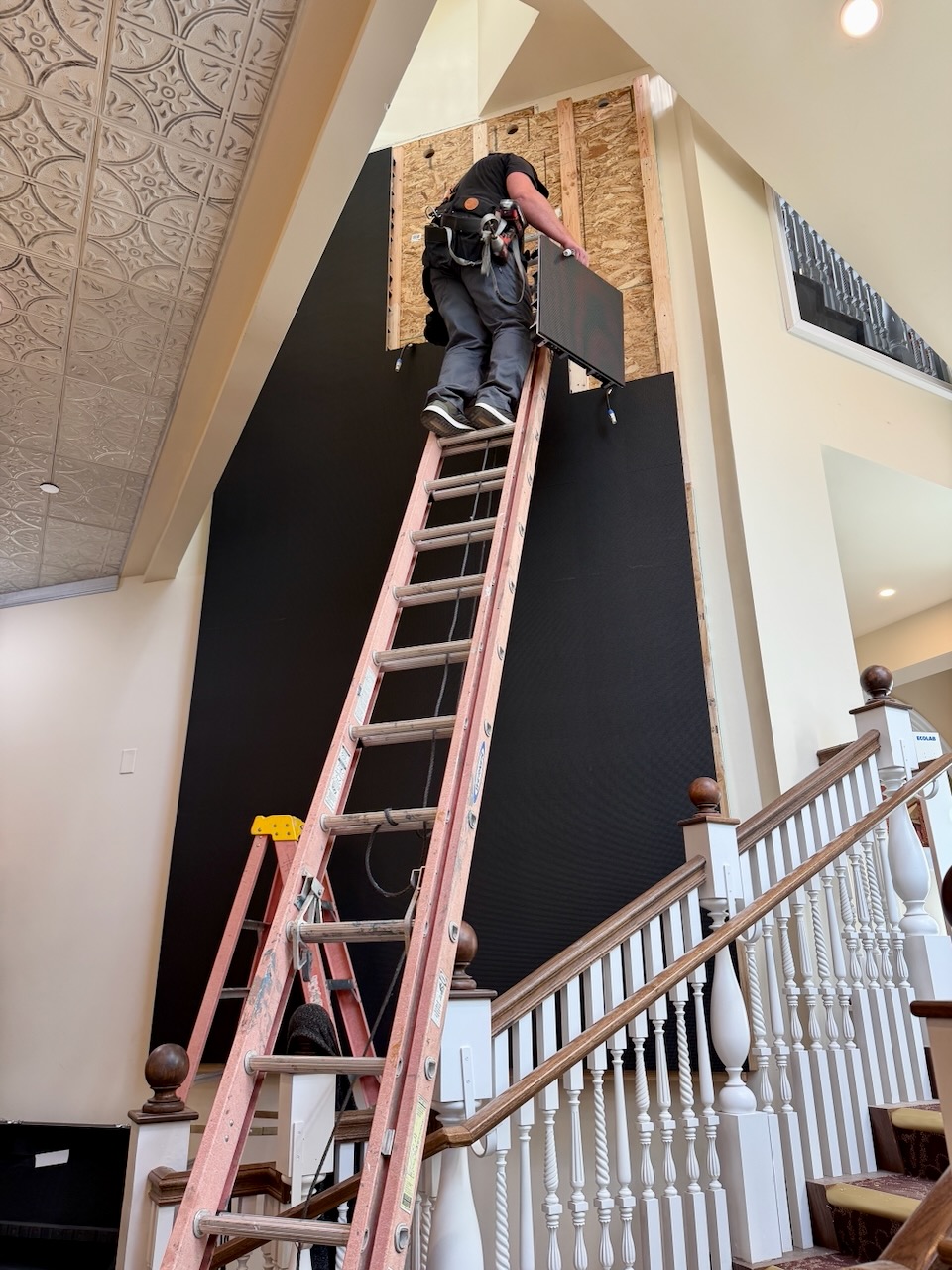 Technician on a 30-foot ladder mounting an LED panel to the studded wall