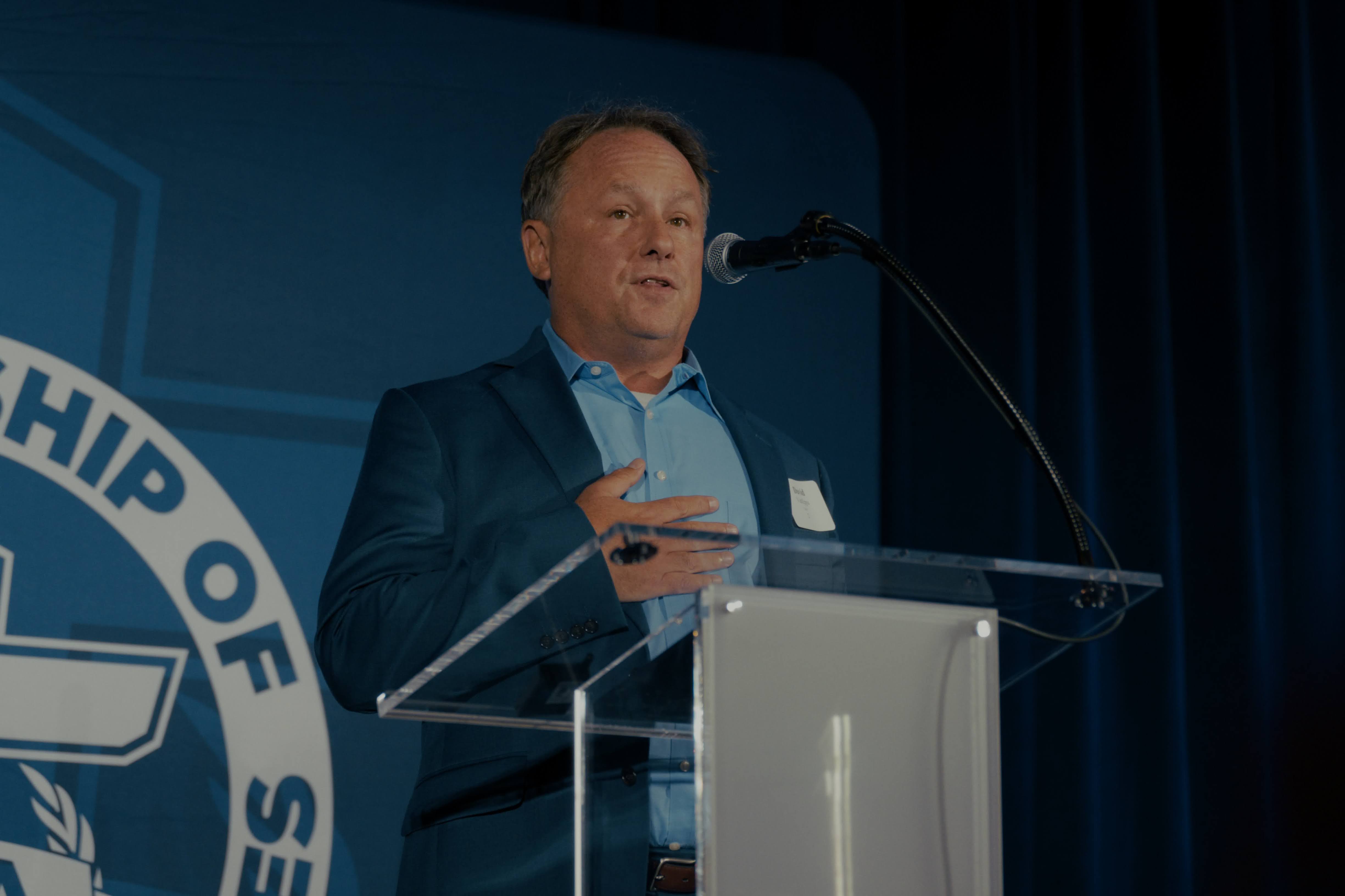 Dave VanEpps speaking at an FCA event behind a clear lectern and microphone.
