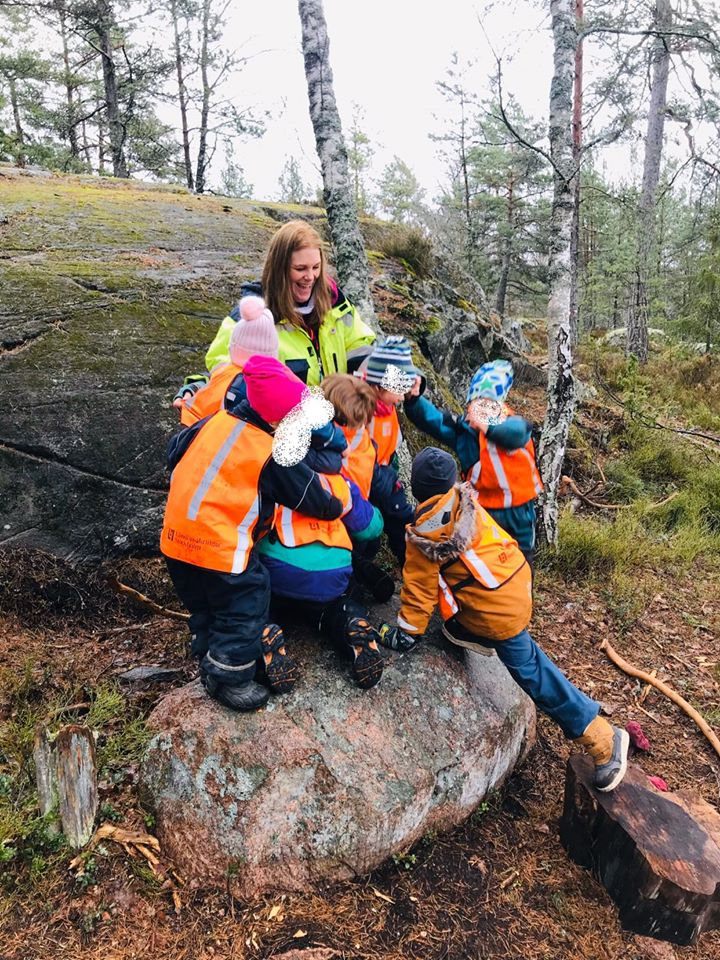 Pedagog och barn i reflexvästar klättrar på ett stort block i skogen