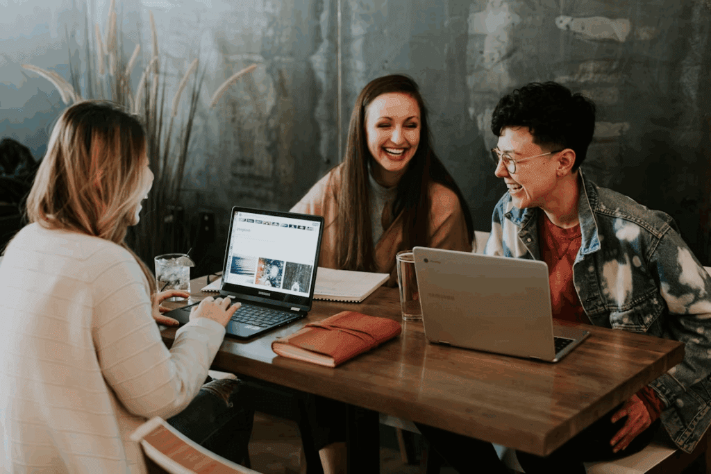 Three co-workers seated around a table, leaning in and laughing together in a bright, relaxed office setting