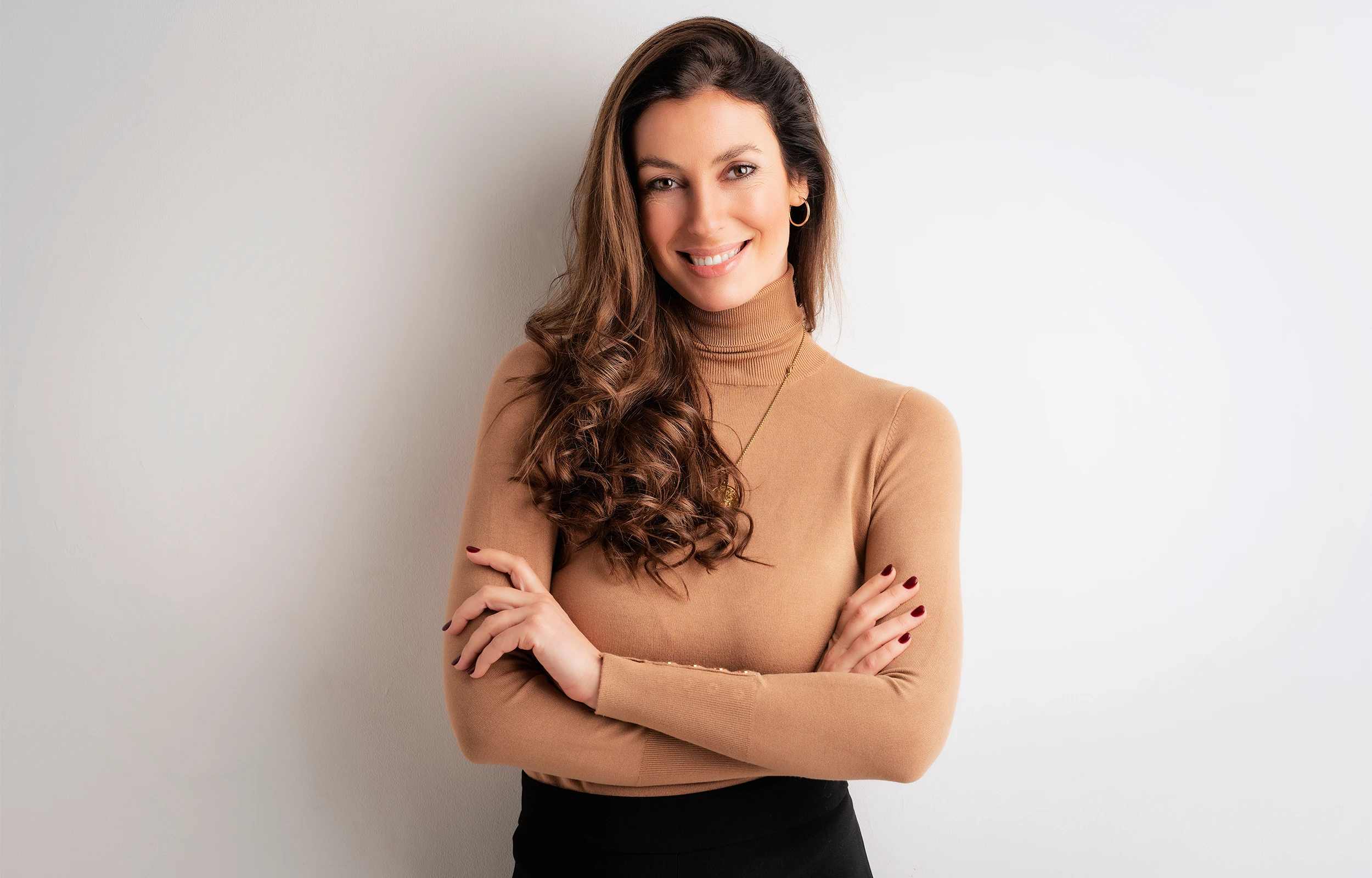 Smiling woman with long curly brown hair wearing a camel turtleneck and black skirt, standing with arms crossed against a white wall.