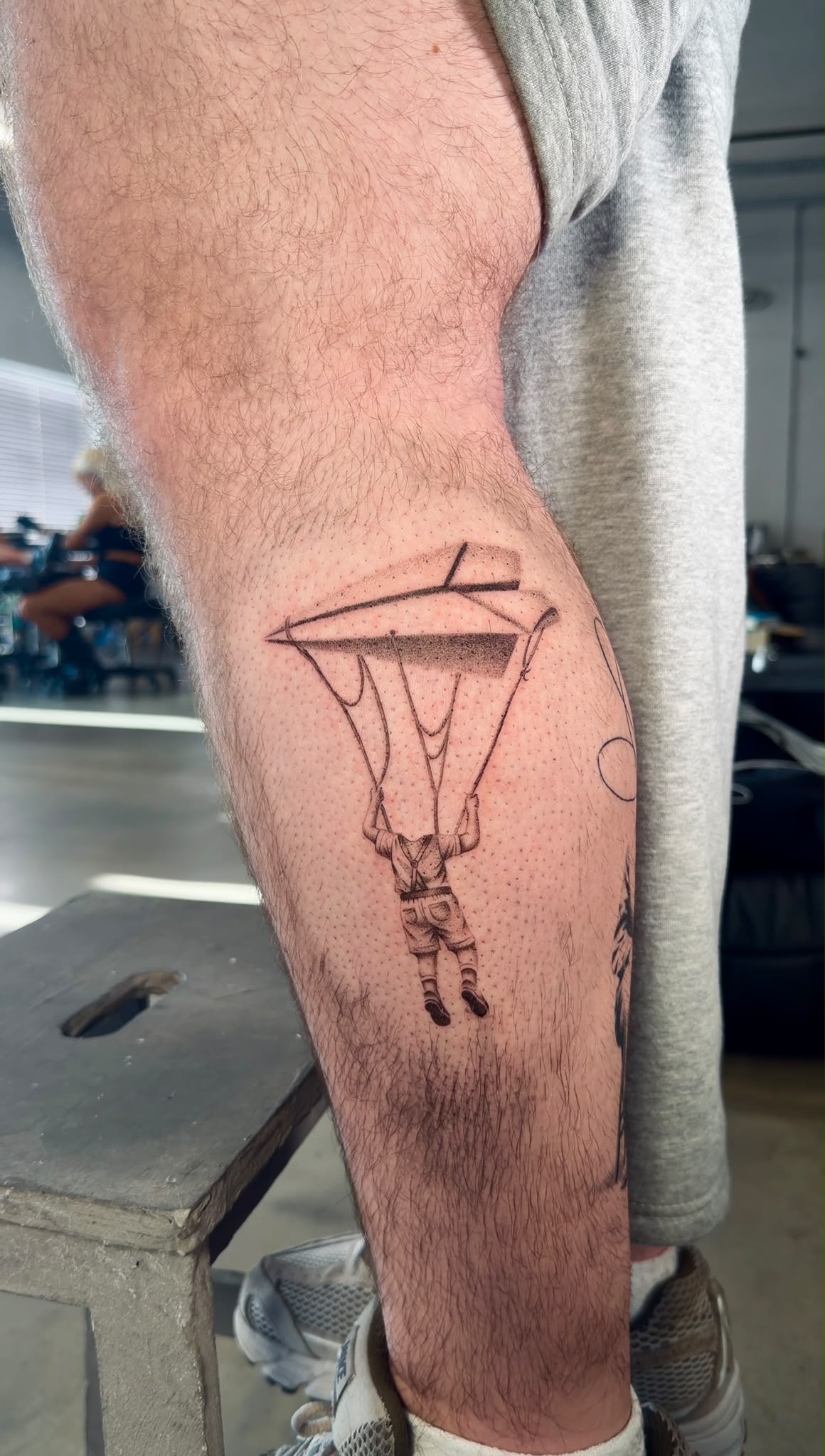 Surreal fine line tattoo of a figure hanging from a paper plane, on the calf, by Tyrese Gilzene at SANS PATRIE/ STUDIO