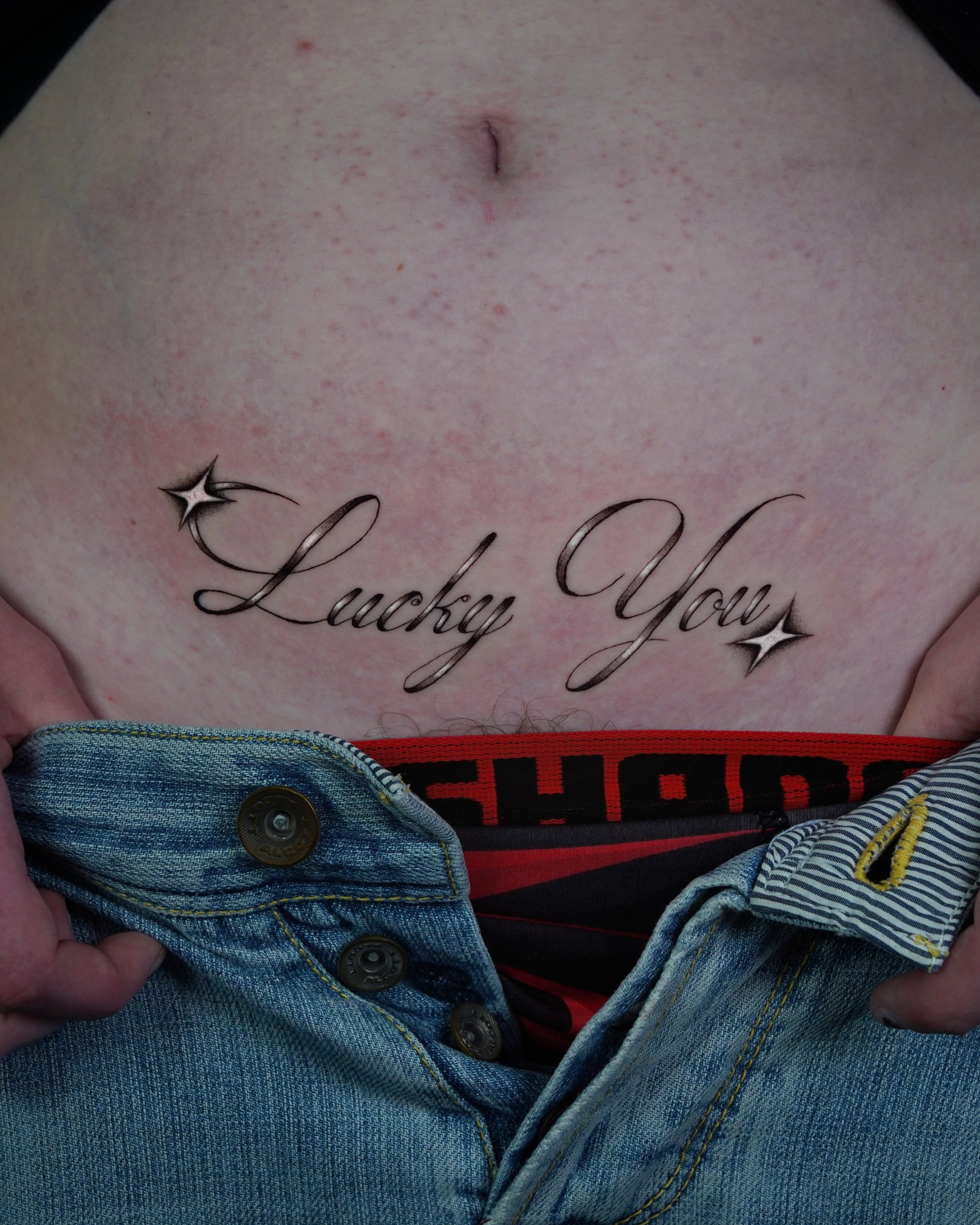 "Lucky You" script lettering stomach tattoo with sparkles by Tommy Burley at SANS PATRIE/ STUDIO