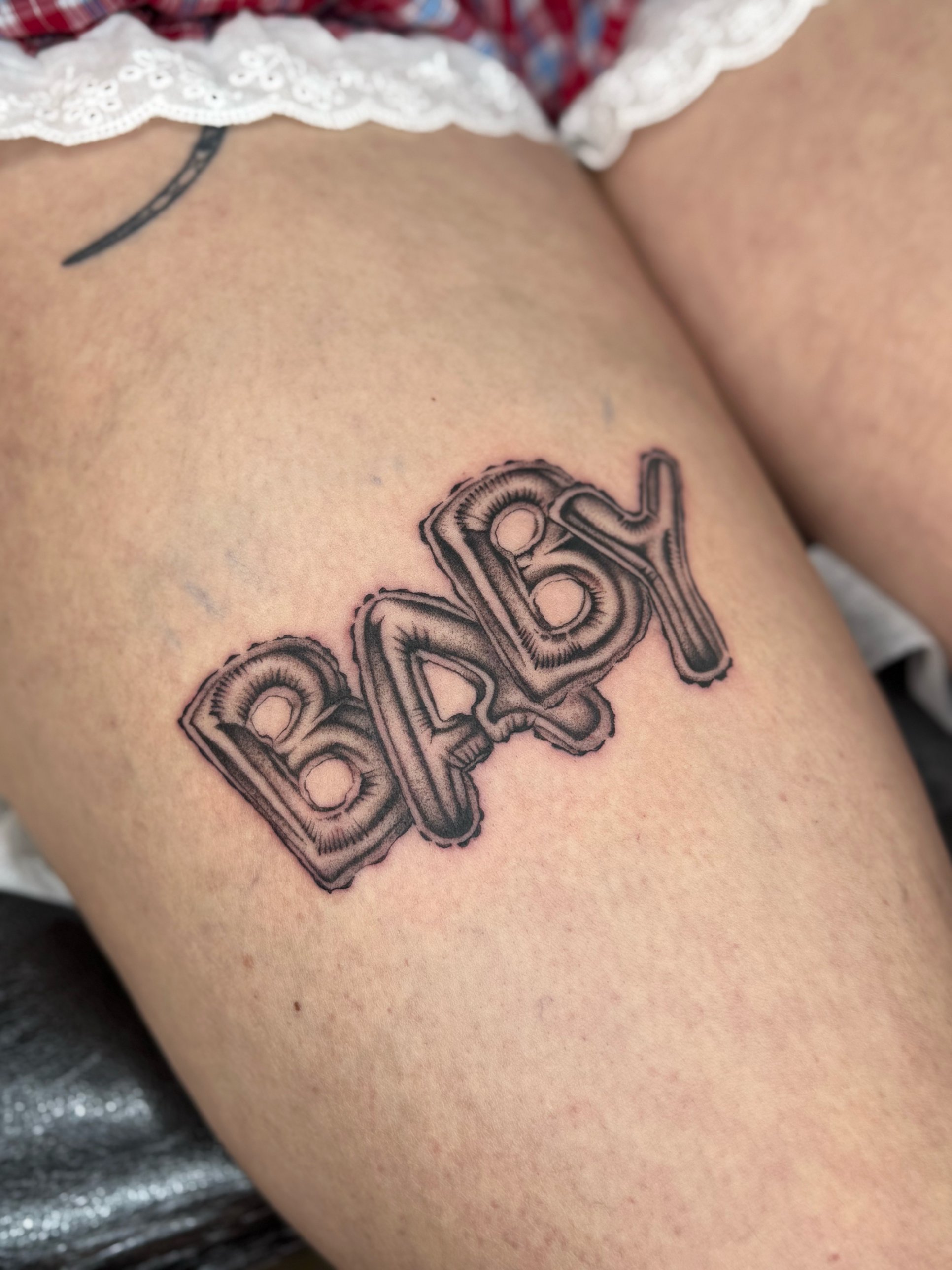 Black and grey foil balloon lettering "BABY" thigh tattoo by Tommy Burley at SANS PATRIE/ STUDIO