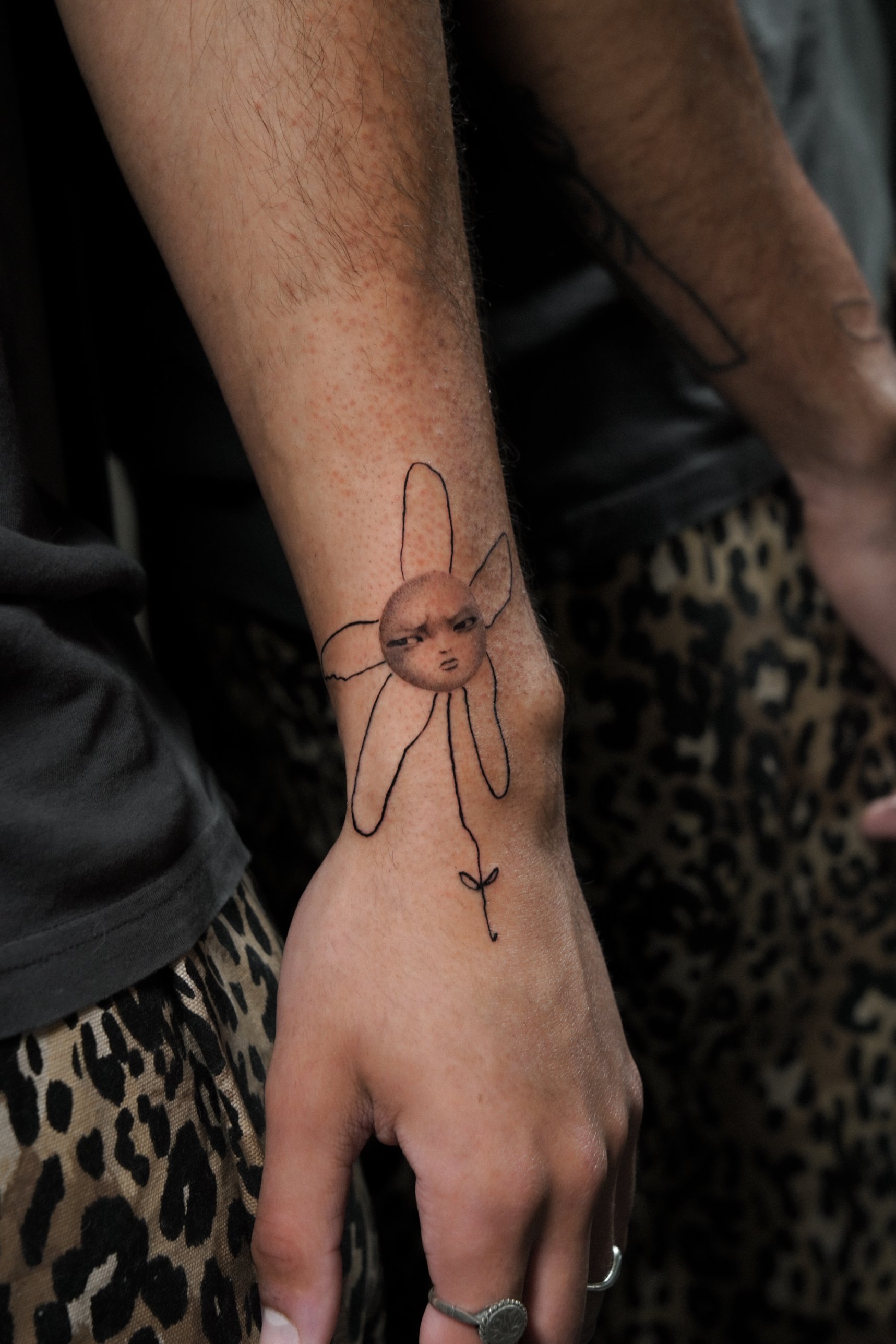 Fine line flower with a surreal face wrist tattoo by Kimberley Verana at SANS PATRIE/ STUDIO
