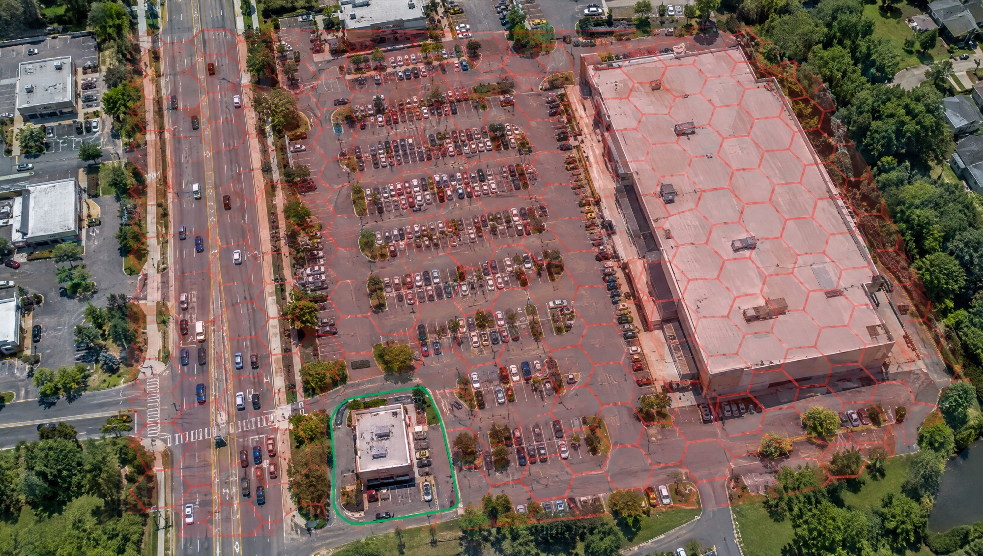 Aerial view of a shopping plaza where a loose red hexagon grid covers the road and parking lot, and a tight green geofence outlines one building