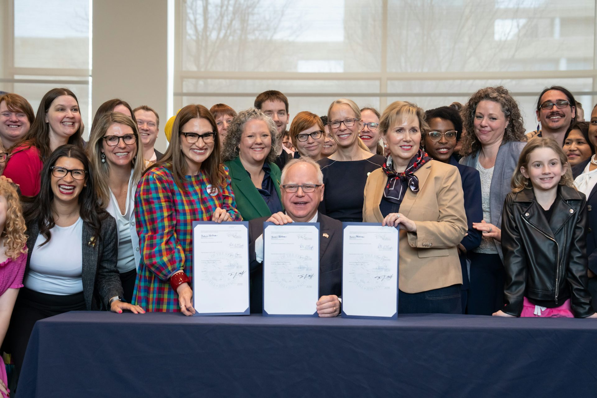 Walz sits smiling at a desk, surrounded by a crowd of smiling people