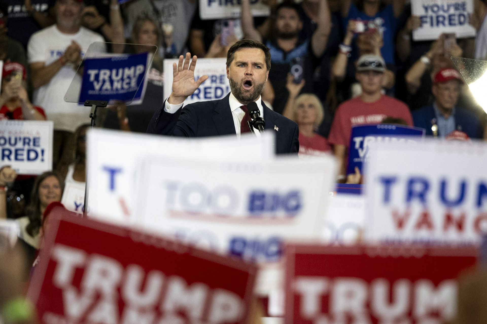 Vance speaks at a rally amid a crowd holding 'Trump-Vance' and 'You're Fired' signs