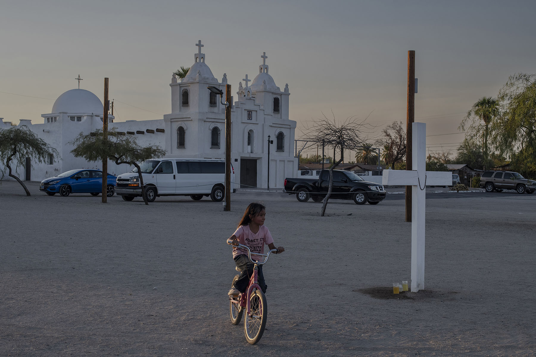 A young girl rides a bicycle in an open square near a white church with two bell towers.