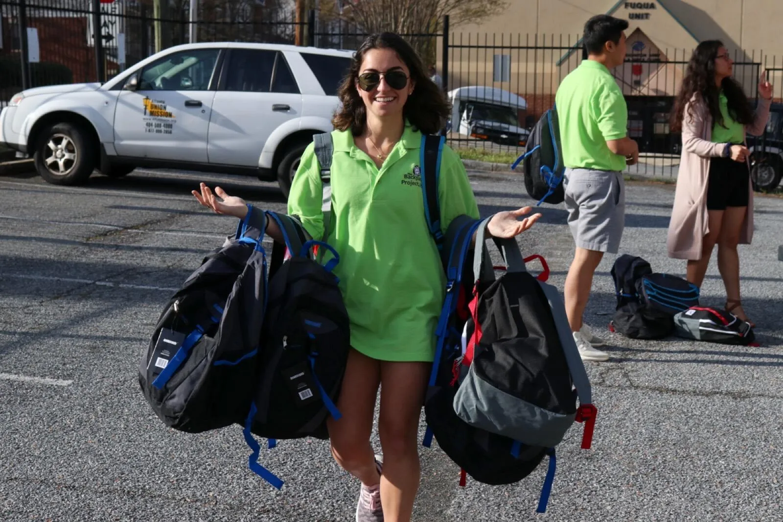 A volunteer in a green Backpack Project polo carries four backpacks across a parking lot.
