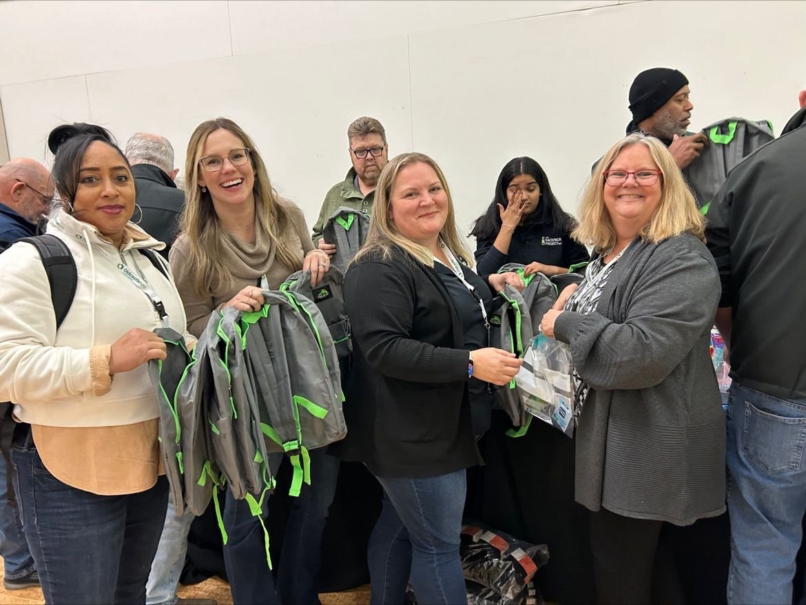Volunteers hold packed grey backpacks with green trim at an Oldcastle packing event.