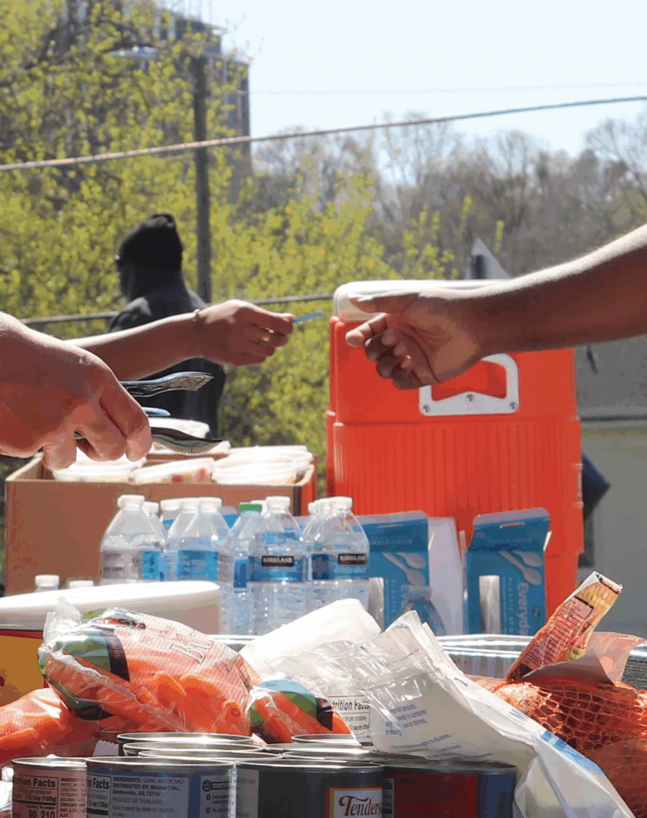 Hands pass bottled water and food from a cooler during an Athens meal distribution.
