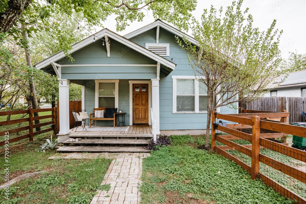 A small blue house with a front porch, a brick walkway and a wooden fence
