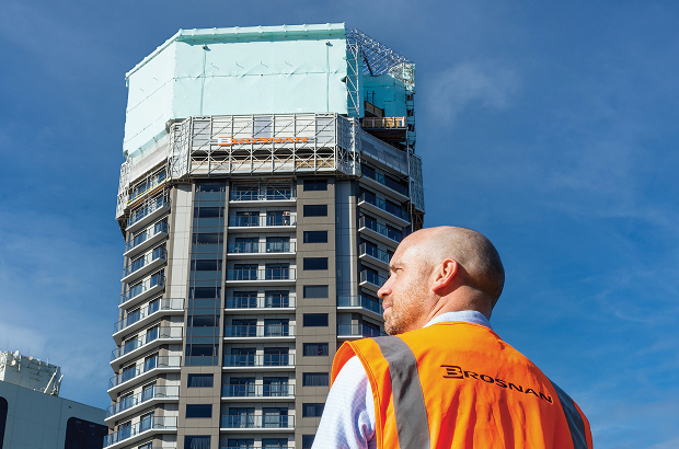 Man in orange safety vest labeled Brosnan looking at a tall building under construction against blue sky.
