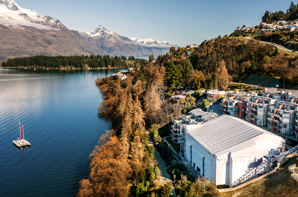 Lake surrounded by trees and buildings with snow-capped mountains in the background on clear day.
