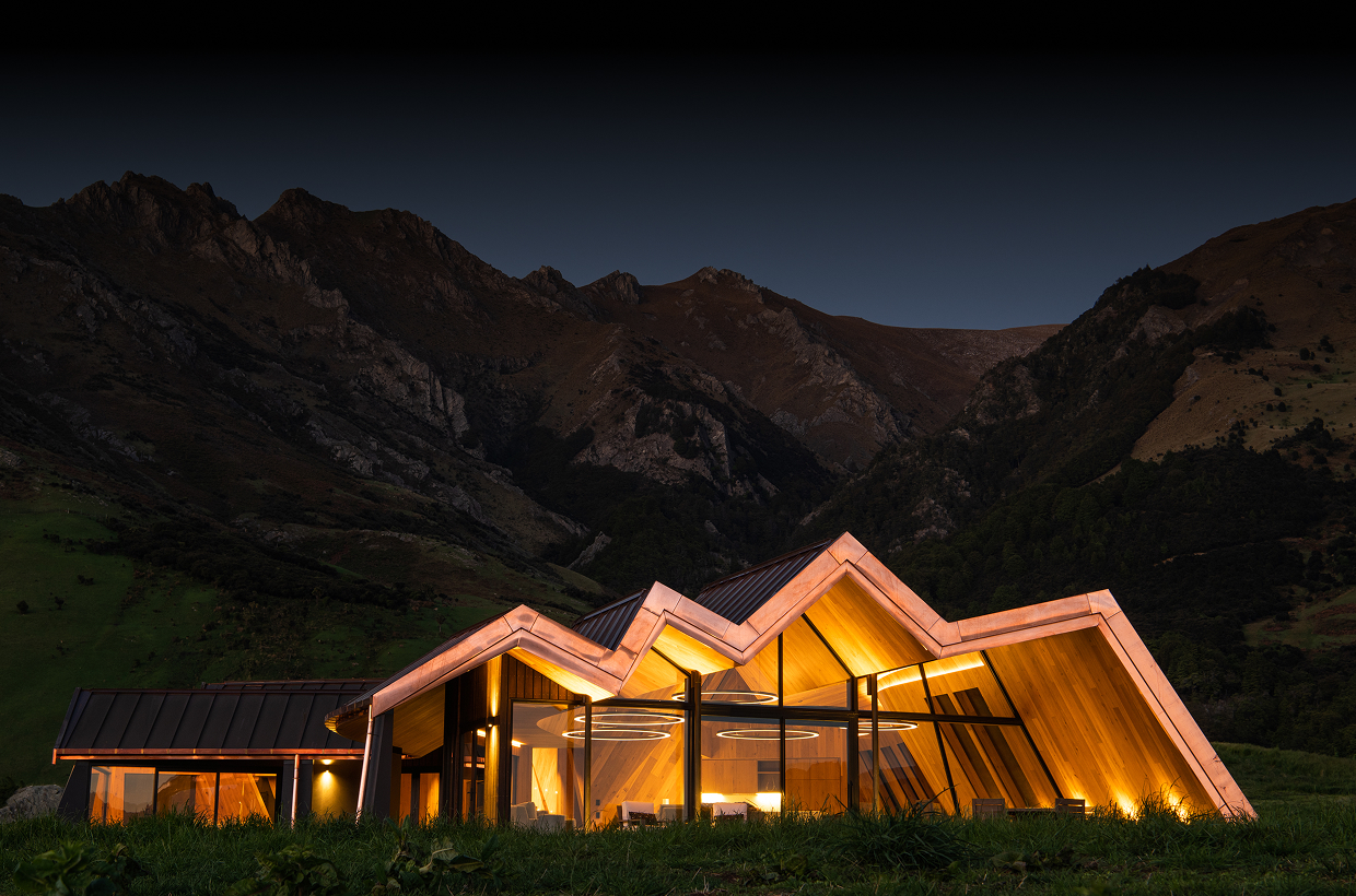 Modern illuminated house with angular roof at night in a grassy valley near steep mountains.