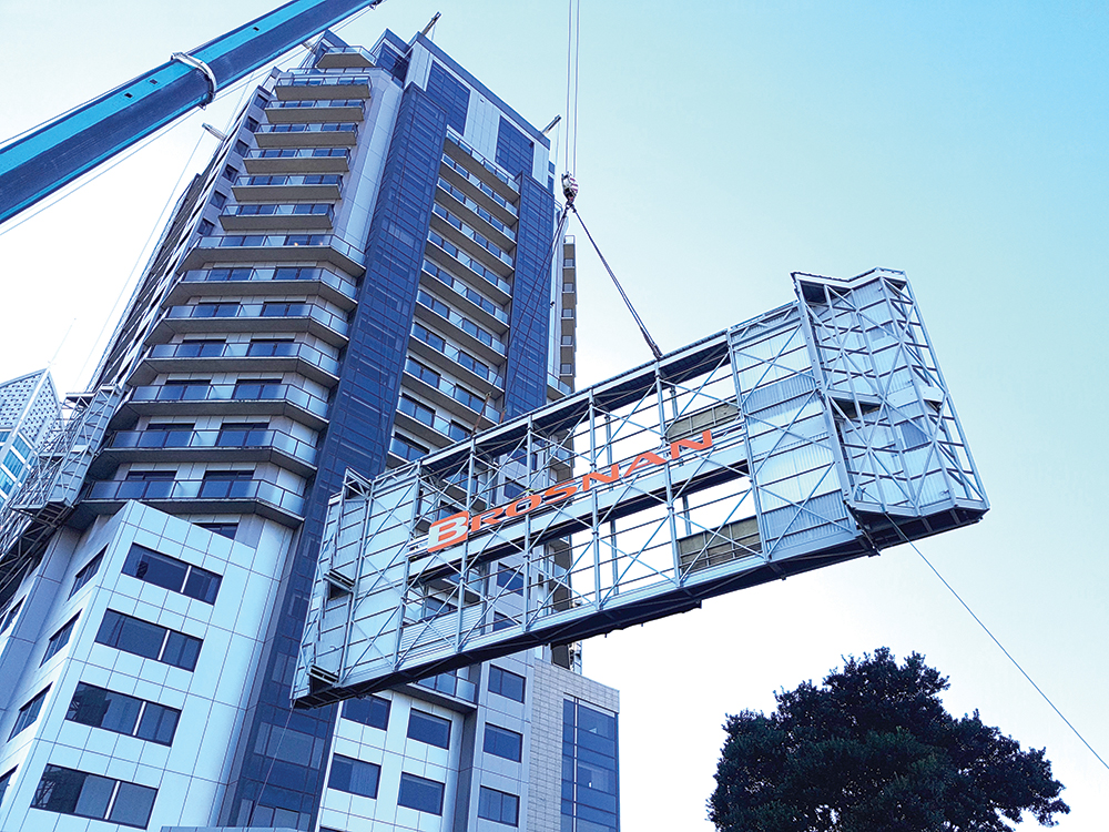 Crane lifting large metal structure with Bro-NaN sign near a high-rise building.