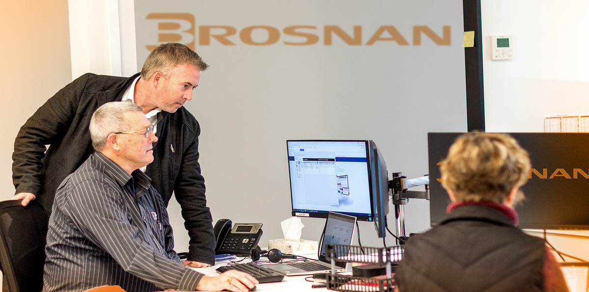 Two men focused on a computer screen in an office with a woman working nearby and Brosnan logo on the wall.