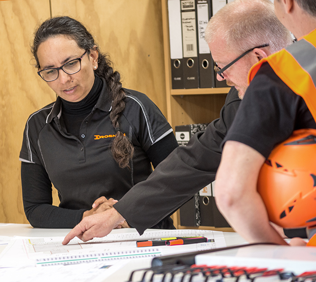 Three coworkers discussing blueprints on a table in an office with folders in the background.