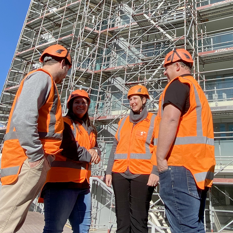 Four construction workers wearing orange safety vests and helmets talking near scaffolding.