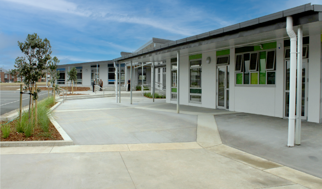 Modern school building with white exterior, covered walkway, and some people near the entrance under a blue sky.