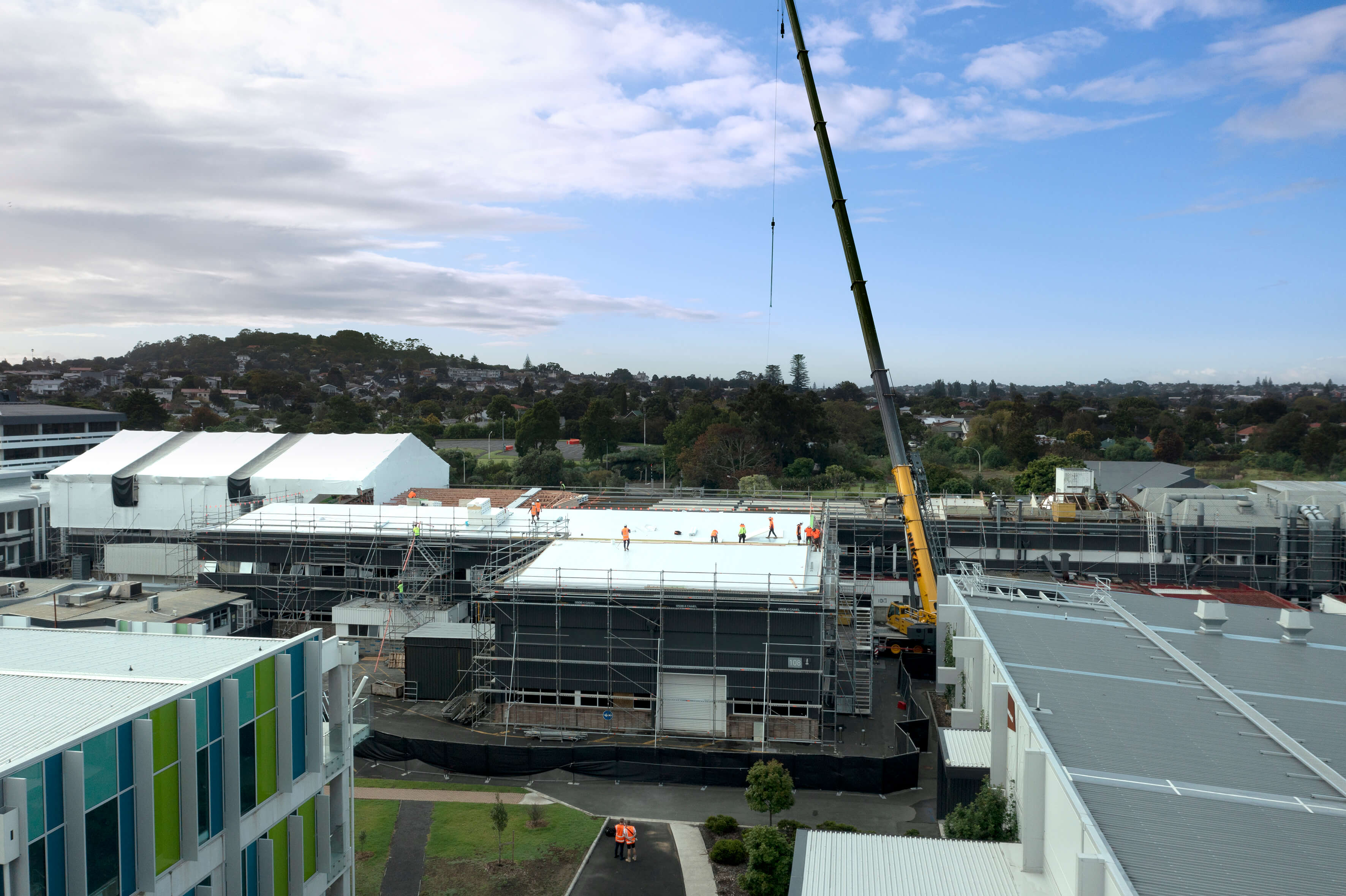 Construction site with workers on a white roof and a large crane lifting materials near buildings and greenery.