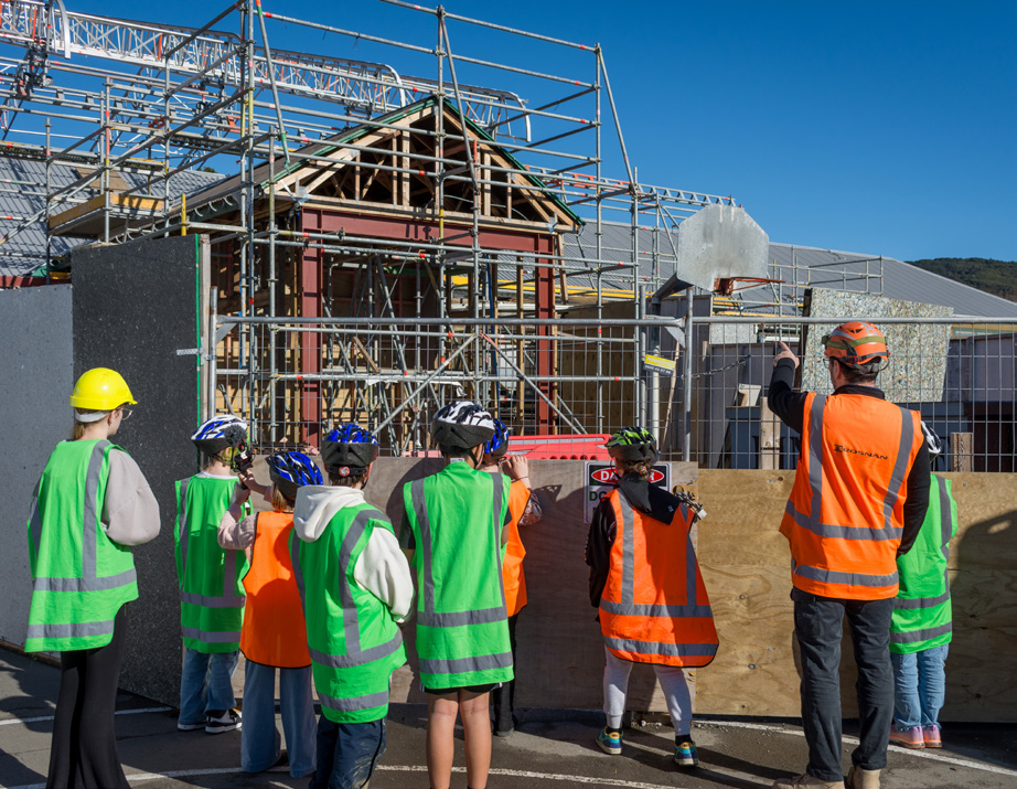 Group of children and an adult in safety vests and helmets looking at a building construction site.