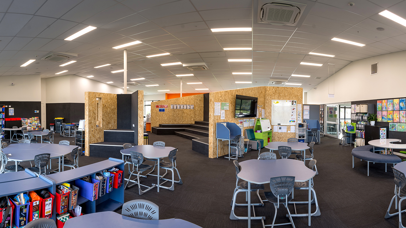 Modern classroom with hexagonal tables, chairs, colorful bins, a TV, and a tiered seating area.
