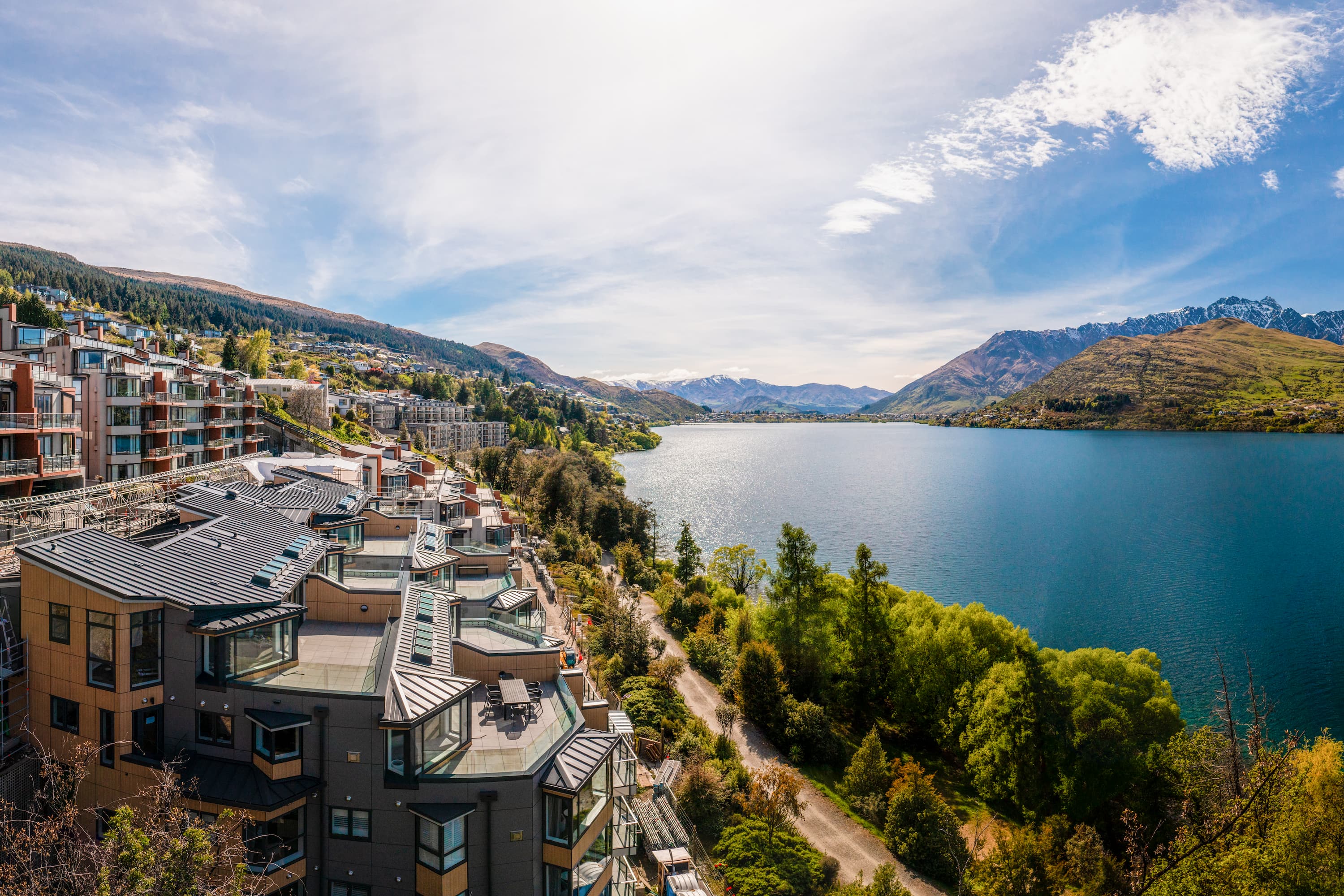 Modern lakeside buildings overlooking a large lake with mountains and trees under a bright sky.