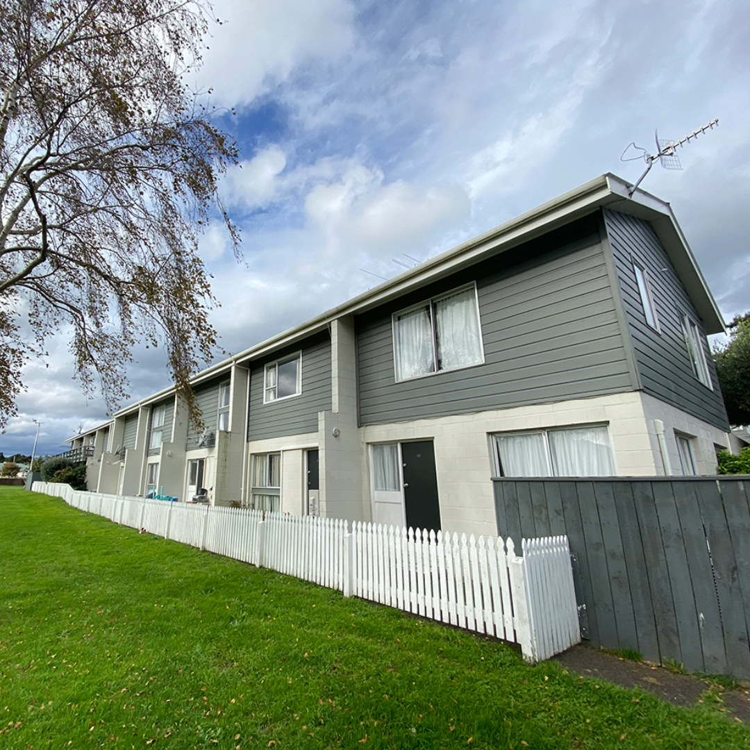 Row of two-story grey and white townhouses with white picket fences and green lawn under a cloudy sky.