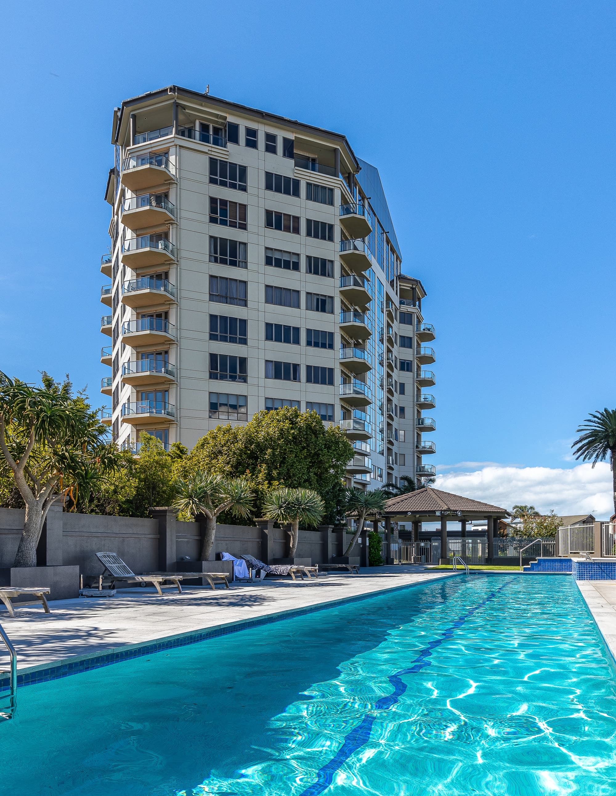 Outdoor swimming pool in front of a multi-story residential building under clear blue sky.