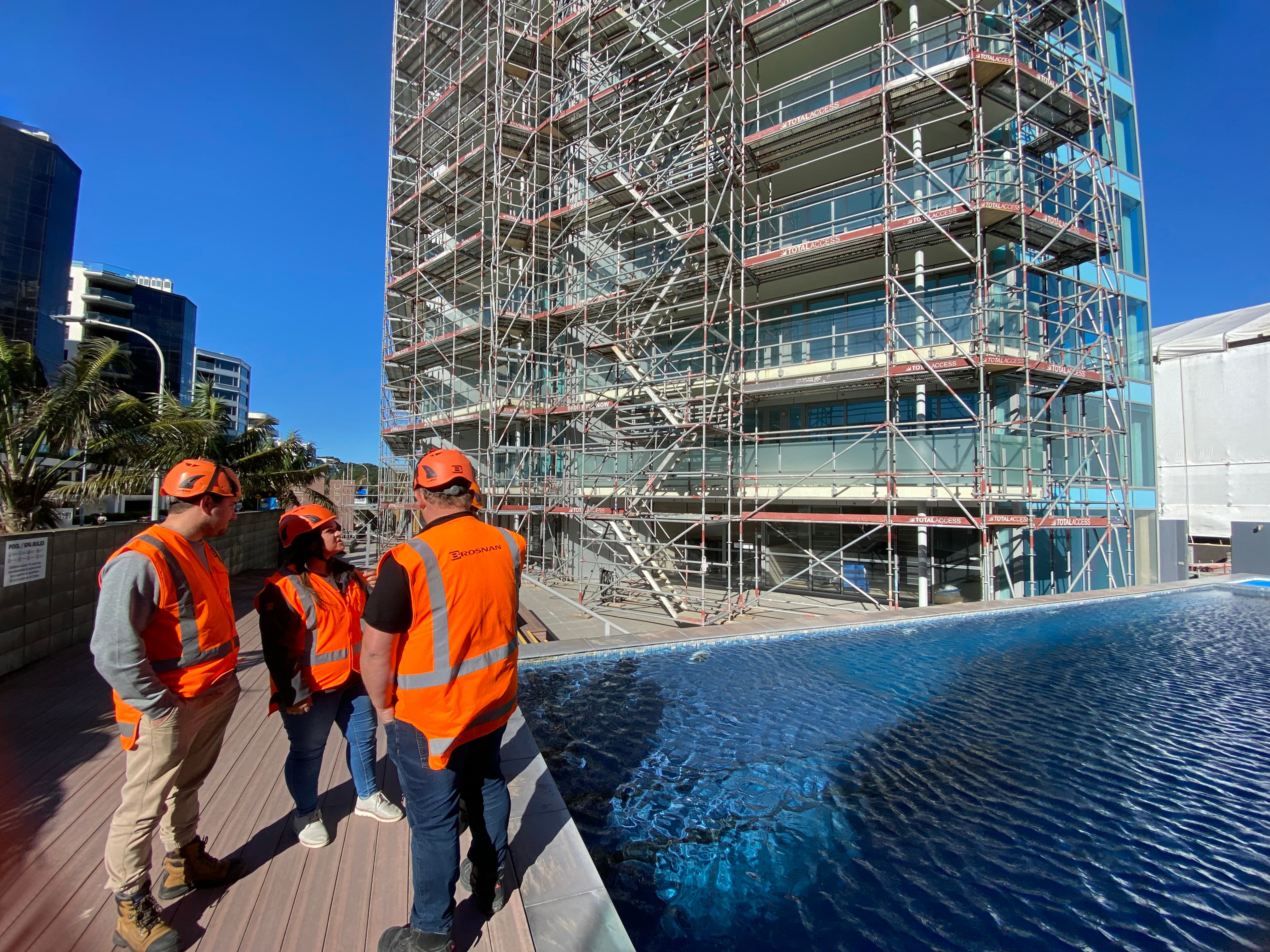 Three construction workers in orange safety gear inspecting building scaffolding near a pool under clear blue sky.