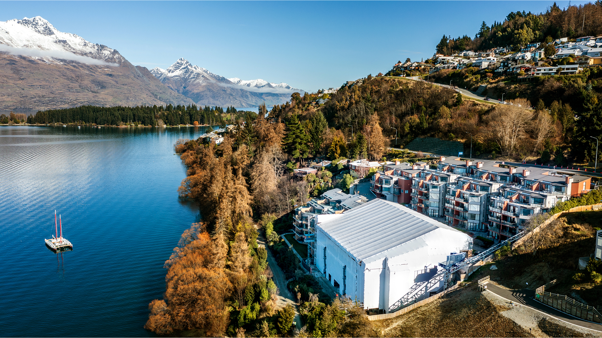 Lake with a sailboat next to autumn trees and buildings at the foot of snow-capped mountains.