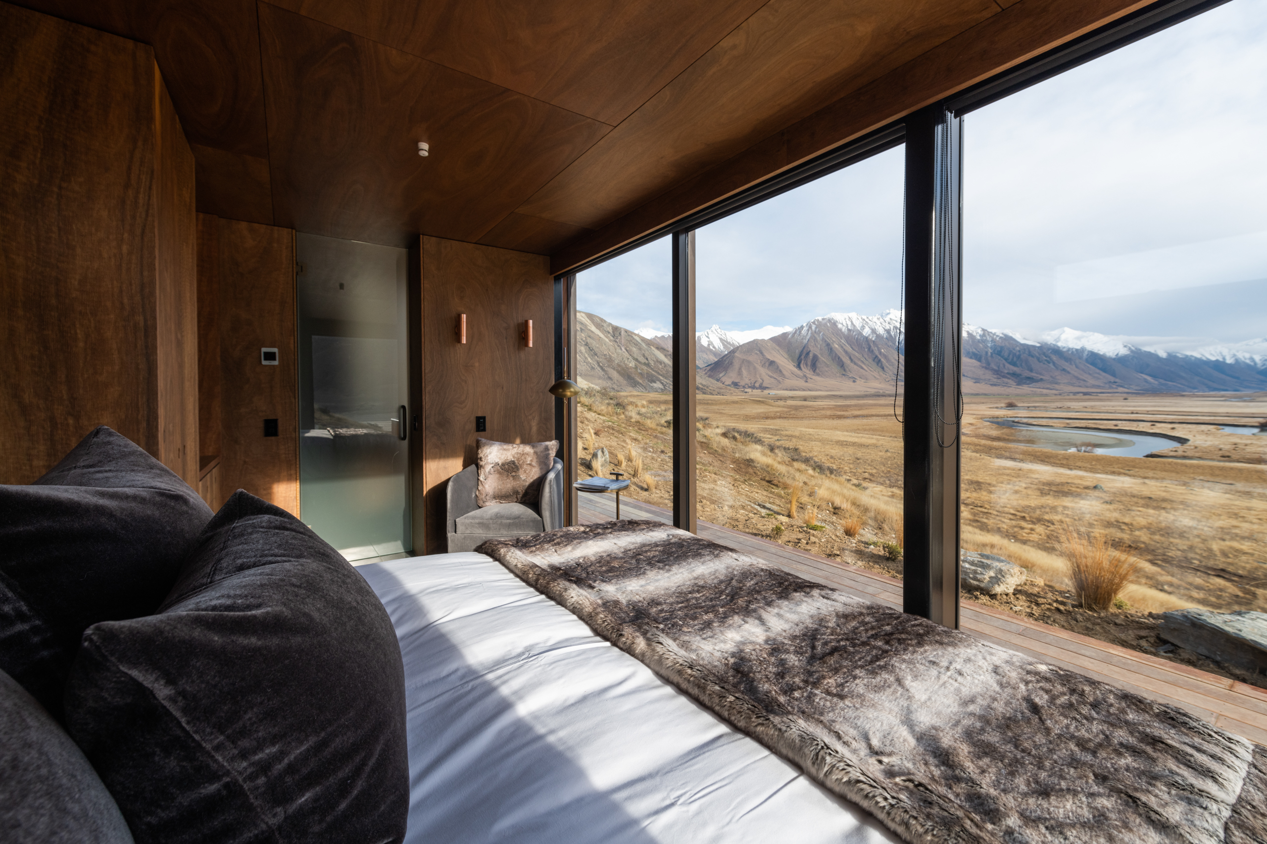 Modern bedroom with large windows overlooking mountains, river, and dry grassland.