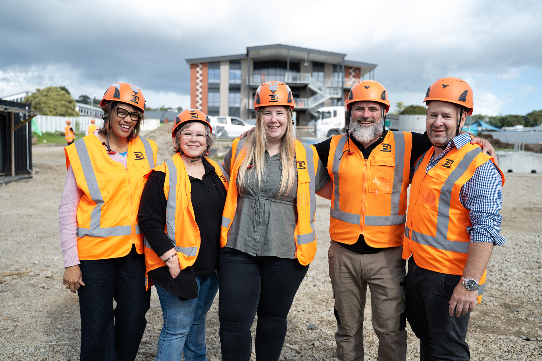 Five construction workers wearing orange safety vests and helmets standing together at a construction site.