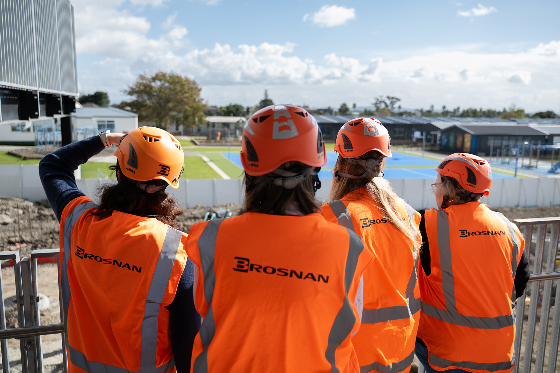 Four people wearing orange Brosnan safety vests and helmets look over a fenced construction site.