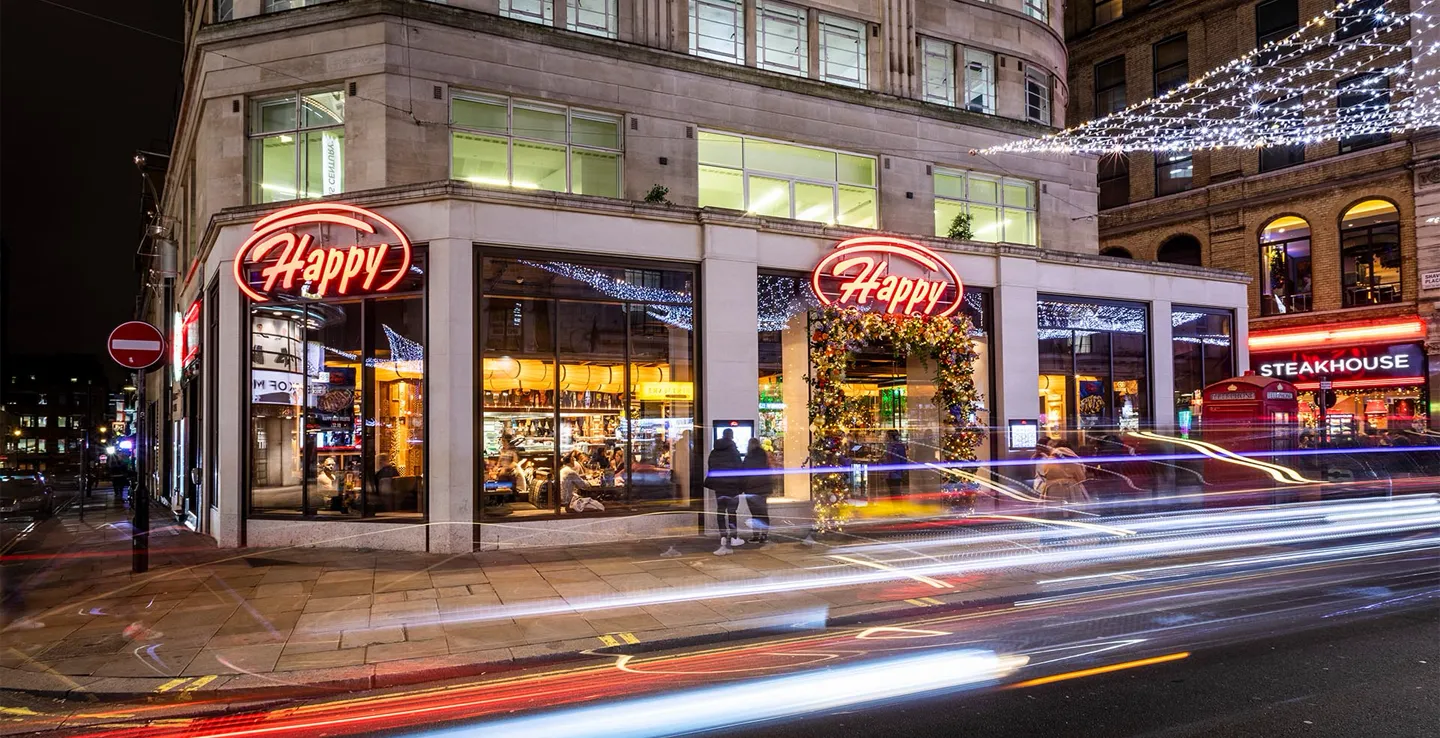 Night view of a corner restaurant with large glass windows and two red neon signs reading 'Happy'; the entrance is decorated with a floral arch; light trails of passing cars blur in front, and a steakhouse with red neon signage and a red telephone booth are visible in the background.