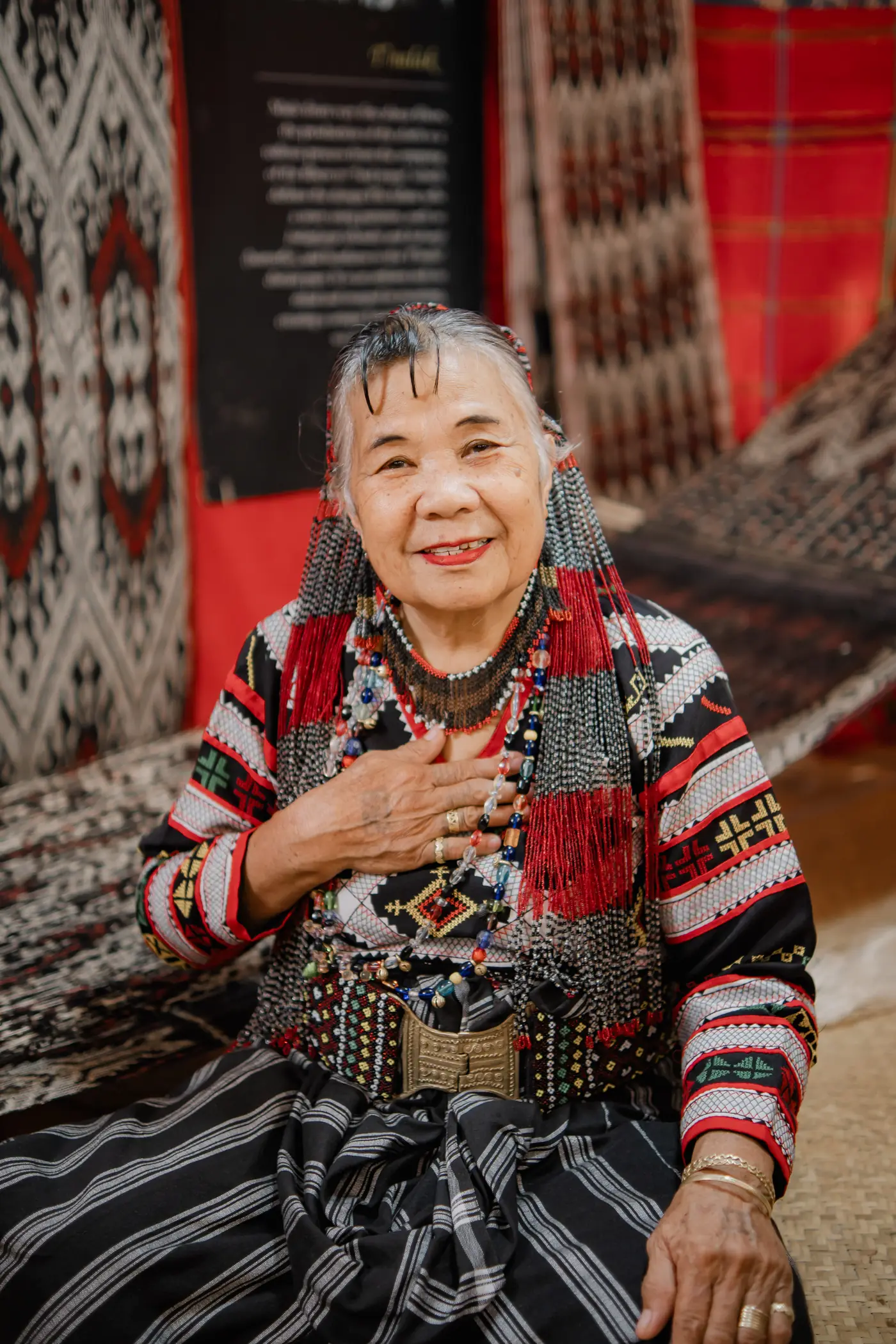 A weaver in traditional dress, smiling, hand on her heart