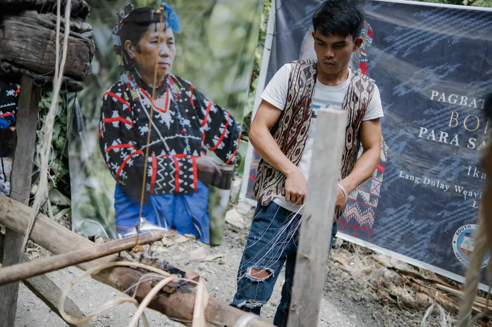 An elder weaver beside a young man working at a loom