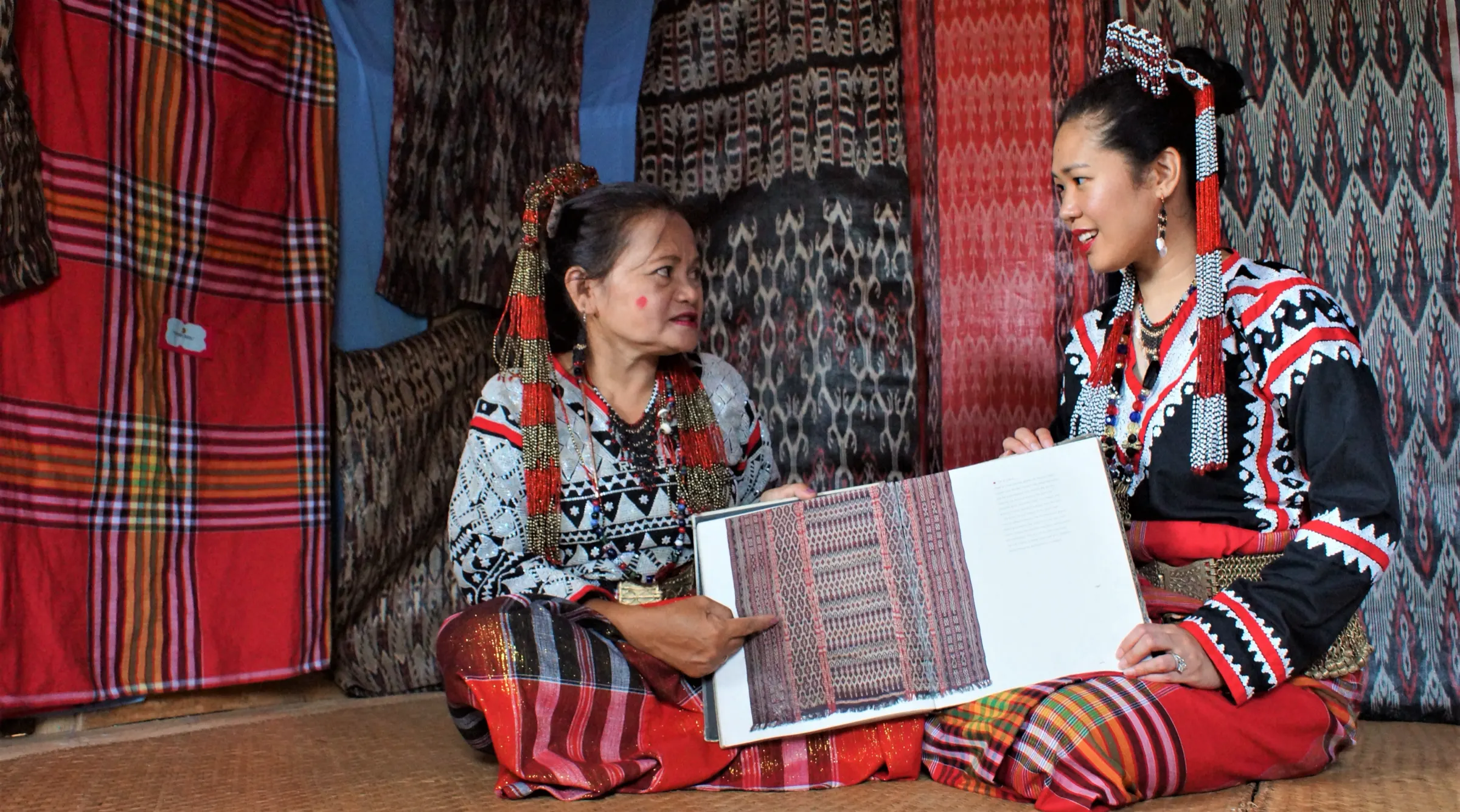 Two women in Tboli dress seated on a woven mat among hanging T’nalak, looking together at a book of textile patterns