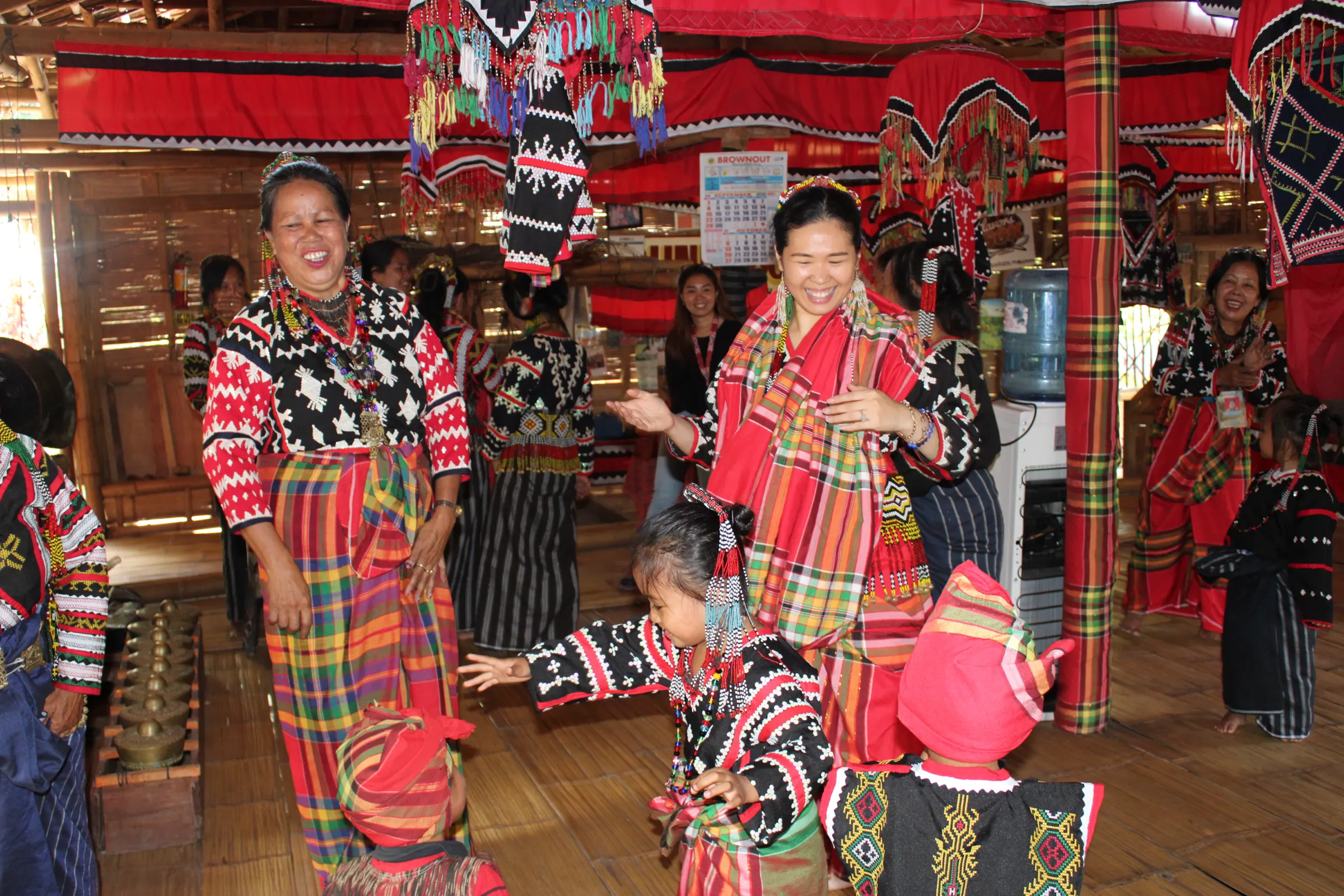 Women and children in Tboli dress dancing and laughing at a gathering, under red canopies hung with beadwork