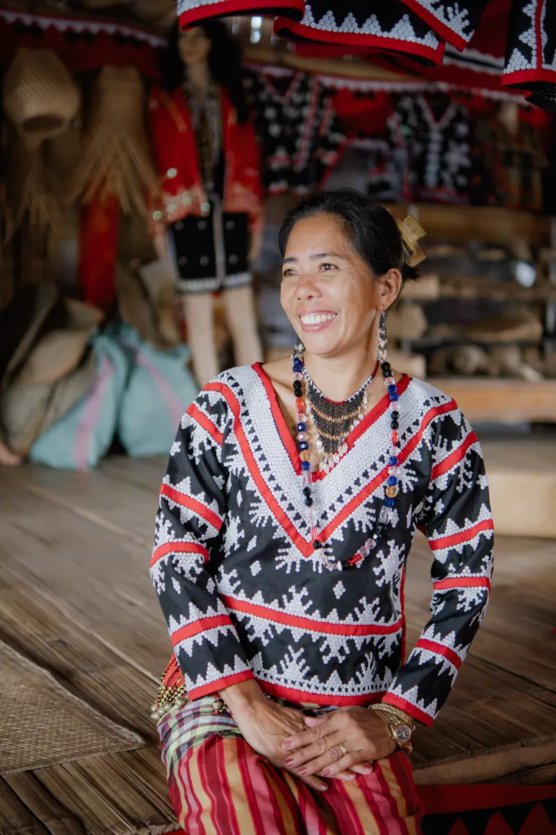 An artisan in Tboli dress seated in a workshop, smiling, with woven goods behind her